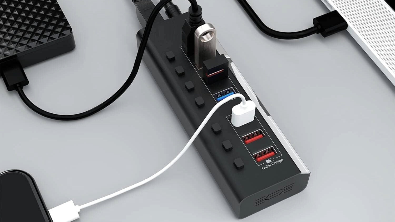 Black multi-port USB hub with power switches, Quick Charge USB ports, and multiple connected devices on a modern desk setup.